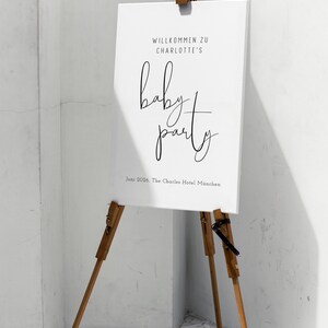 Puede incluir: Un cartel blanco con texto negro en un caballete de madera. El cartel dice "Willkommen zu Charlotte's baby party Juni 2026, The Charles Hotel Munchen".