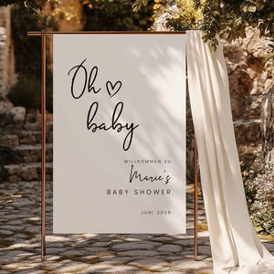 Puede incluir: Un cartel blanco con texto negro que dice "Oh baby" con un s&iacute;mbolo de coraz&oacute;n. El cartel tambi&eacute;n dice "Willkommen zu Marie's Baby Shower Juni 2026". El cartel est&aacute; apoyado en un soporte dorado frente a una cortina blanca.