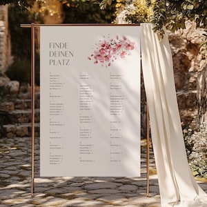 May include: A rectangular wedding seating chart sign on a copper-colored metal stand, featuring the German text FINDE DEINEN PLATZ, a pink floral illustration, and a list of names organized alphabetically with corresponding table numbers.