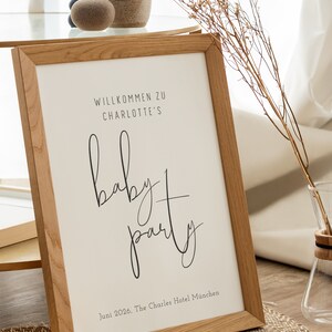 Puede incluir: Un cartel enmarcado con un marco de madera y fondo blanco. El cartel dice "Willkommen zu Charlotte's Baby Party" y "Juni 2026, The Charles Hotel M&uuml;nchen".