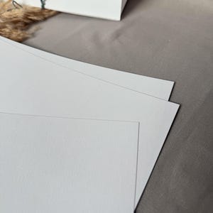 May include: Three white blank cards stacked on top of each other on a light brown surface.