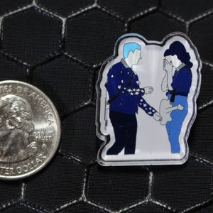 May include: A blue and white enamel pin featuring a cartoon couple. The man is wearing a blue jacket with white stars and the woman is wearing a blue shirt and jeans. The woman is crying and the man is holding her hand.