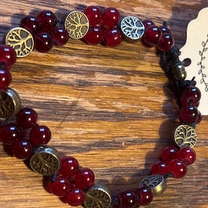 May include: A beaded bracelet featuring deep red, round beads and circular metal charms with a tree design. The bracelet is strung on a black cord and rests on a wooden surface.