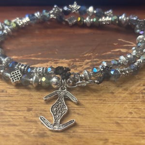 May include: A silver bracelet with two strands of iridescent beads and decorative metal elements. The bracelet has an anchor charm and a star-shaped clasp. The beads are a mix of colours, including blue, green, and silver.