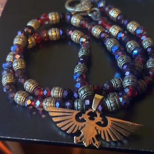 May include: A necklace featuring deep red faceted crystal beads alternating with antique brass-toned cylindrical spacers. The piece includes a central pendant shaped like a stylized eagle with outstretched wings, finished in a matching brass tone.
