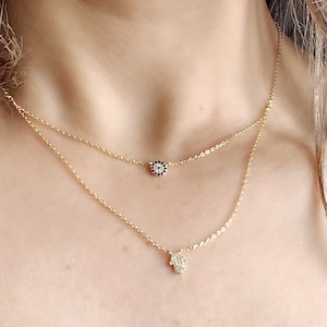 Könnte beinhalten: Zwei goldene Halsketten mit einem kleinen bösen Augen-Anhänger und einem Hamsa-Hand-Anhänger. Beide Anhänger sind mit kleinen weißen Steinen besetzt.