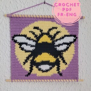 Könnte beinhalten: Ein quadratischer Wandbehang aus Häkelarbeit mit einem Hummelmotiv in Schwarz, Gelb und Weiß auf violettem Grund. Das Stück hängt an einem Holzstab mit einer Aufhängung aus violettem Garn. Der Text auf dem Bild lautet CROCHET PDF FR-ENG.