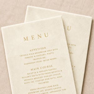 May include: Two off-white rectangular menu cards with gold lettering. The text lists sections for Appetiser, Main Course, and Dessert, including items like Truffle Wild Mushroom Soup, Ricotta & Spinach Ravioli, Herb-Crusted Lamb Rack, and Classic Tiramisu.