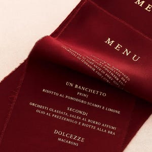 May include: A deep burgundy fabric menu with frayed edges and white printed text. The menu features the headings MENU, UN BANCHETTO, PRIMI, SECONDI, and DOLCEZZE, followed by Italian food items including Risotto al Pomodoro Scampi e Limone and Macarons.