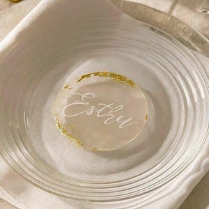 May include: A circular, translucent place card with a smooth, frosted finish and irregular gold leaf edges. The name Esther is written in white cursive script across the center of the disc, which is resting on a white napkin atop a clear glass plate.