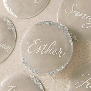May include: A collection of circular, semi-translucent gray acrylic discs featuring white cursive names printed on the surface. Each disc is accented with a textured silver leaf edge detail along a portion of the perimeter, creating a decorative effect.