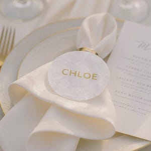 Op de afbeelding: Een rond wit gemarmerd acryl naamkaartje met de naam CHLOE in gouden letters, bevestigd aan een gouden metalen servetring die een gevouwen wit stoffen servet vasthoudt op een wit bord met een gouden rand en een geprinte menukaart.