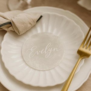 May include: A white scalloped plate holding a translucent circular place card with the name Evelyn written in white script. A gold-toned fork rests on the right side of the plate, accompanied by a beige fabric napkin folded on the left side.
