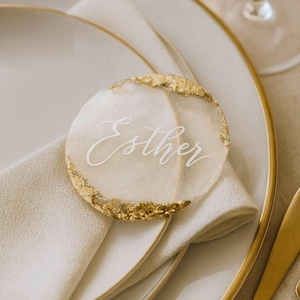 May include: A circular, translucent place card with a smooth finish, featuring the name Esther written in white script. The edges of the disc are accented with textured gold leaf detailing, creating a decorative border around the perimeter.