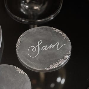 May include: A circular, translucent resin coaster featuring a cloudy white finish with silver glitter accents along the edges. The name Sam is written in the center in a white, cursive script font. A second, matching coaster is partially visible below.