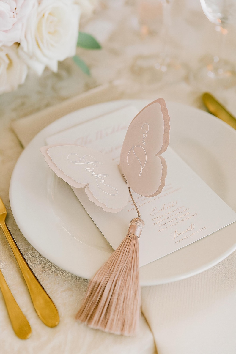 May include: A butterfly-shaped place card with a tassel, resting on a white plate. The place card is light pink with white script. A menu card is visible beneath the place card. Gold silverware and white flowers are in the background.