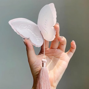 May include: A light pink butterfly-shaped ornament with the name "Samantha" and the word "Loren" written on the wings. The ornament has a matching pink tassel attached to it.