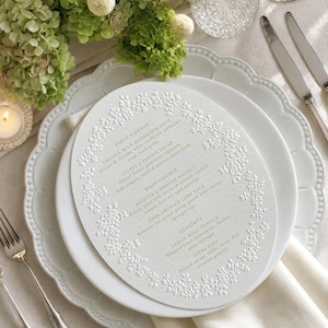 May include: An oval-shaped white menu card with embossed floral borders and gold text. The menu lists First Course, Main Course, and Desserts, including items like Truffle Wild Mushroom Soup, Ricotta Spinach Ravioli, Herb-Crusted Lamb Rack, and Opera Cake.