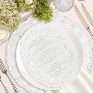 Może przedstawiać: Eleganckie nakrycie stołu z białymi talerzami, sztućcami i menu. Menu ma kwiatowy wzór i listę dań, w tym zupę z truflami i deser waniliowo-bazyliowo-cytrynowy. Zielono-biała kompozycja kwiatowa.