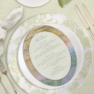 Könnte beinhalten: Eine elegant gedeckte Tafel mit einem Teller mit Blumenmuster, einem weißen Teller und einer Menükarte mit einem Gemälde im Monet-Stil. Die Menükarte lautet „Zum Abendessen“ und listet Vorspeisen, Hauptgerichte und Desserts auf. Ein grünes Namensschild und Besteck vervollständigen das Set.