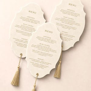 May include: Three off-white, scalloped-edge menu cards with gold-coloured text and gold tassels attached at the bottom. Each card lists appetisers, main courses, and desserts, including truffle mushroom soup, ricotta ravioli, and lamb rack.