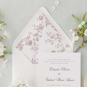Puede incluir: Un conjunto de invitaciones de boda con una tarjeta de invitación blanca con una escritura elegante, un sobre con un forro floral y ramitas de flores blancas. El texto de la invitación dice "Claudia Olivia to Gabriel Dean Louise."