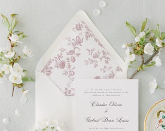 May include: A white wedding invitation card with black serif text reading Claudia Olivia to Gabriel Dean Louise, placed inside a matching envelope with a mauve floral patterned liner, surrounded by white cherry blossom branches.