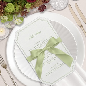 Puede incluir: Elegante mesa con plato blanco, tarjeta de menú y cubertería. La tarjeta de menú, atada con una cinta verde claro, muestra el texto "The Menu". Un arreglo floral con flores rojas y verdes añade un toque de color.