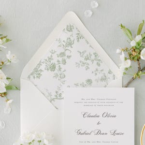 May include: A wedding invitation suite. The white invitation card has elegant script, and is accompanied by an open envelope with a floral patterned lining, and sprigs of white flowers. The invitation reads "Claudia Olivia to Gabriel Dean Louise."