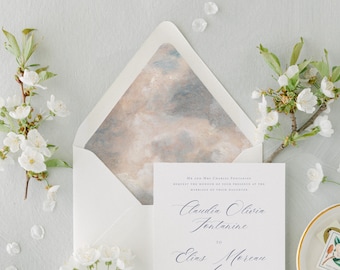 May include: A white wedding invitation card with black serif and script text reading Claudia Olivia Fontanine to Elias Moreau, placed inside an envelope with a marbled beige and grey interior liner, flanked by two branches of white cherry blossoms.