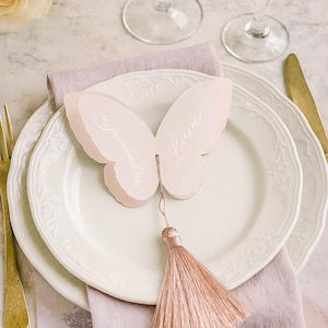 May include: A butterfly-shaped place card with a tassel, resting on a white plate with a floral design. The card is light pink with the names "Samantha" and "Loren" written on it. A gold fork and knife are visible.