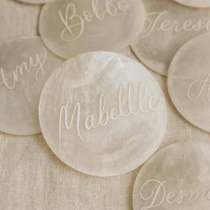 Può includere: Primo piano di diversi segnaposto rotondi bianco sporco con nomi scritti in elegante corsivo bianco. I nomi includono "Cherry", "Bobbo", "Teresa", "Amy", "Mabelle", "A" e "Desme". I segnaposto sono disposti su una superficie beige chiaro strutturata.