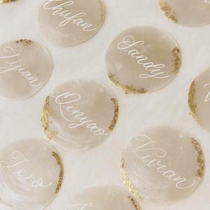 Może przedstawiać: Okrągłe winietki z imionami napisanymi elegancką białą czcionką. Każda karta ma złoty brokatowy brzeg. Karty są ułożone na jasnej powierzchni, odpowiednie na wesela lub specjalne wydarzenia.