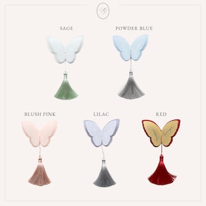 May include: A collection of butterfly-shaped ornaments in various colors: sage, powder blue, blush pink, lilac, and red. Each butterfly has a tassel hanging below. The word "Jacqueline" is written on the red butterfly.