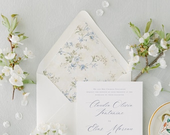 Könnte beinhalten: Eine weiße Hochzeitseinladungskarte mit schwarzem Serif- und Schreibschrifttext, der Claudia Olivia Fontanine to Elias Moreau lautet, in einem Umschlag mit floralem Innenfutter mit blauen und grünen botanischen Illustrationen auf cremefarbenem Papier.