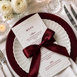 May include: A white rectangular menu card with embossed floral borders and dark red text, featuring a deep burgundy satin ribbon tied in a bow across the center. The menu lists courses including Truffle Wild Mushroom Soup, Citrus and Fennel Salad, and Lemon Basil Vanilla.