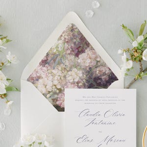 May include: A white envelope with a floral design on the inside flap. The envelope is addressed to Claudia Olivia Fontaine and Elias Moreau. The text "Mr. and Mrs. Charles Fontaine request the honour of your presence at the marriage of their daughter" is printed on the envelope.