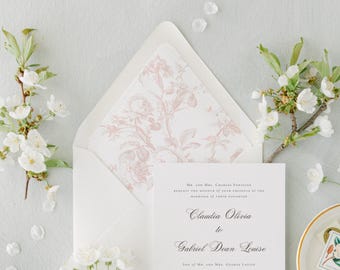 May include: A white wedding invitation card with black serif and script text reading Claudia Olivia to Gabriel Dean Louise, tucked into an envelope with a pale pink floral patterned liner, surrounded by white cherry blossom branches.