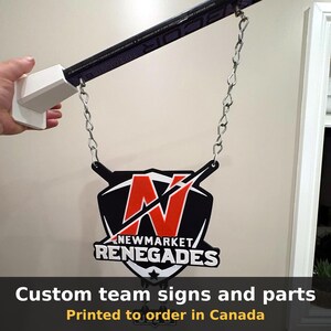 Hand holding a black and white Newmarket Renegades team sign printed in 3D and hanging from a chain, with the text Custom team signs and parts, printed to order in Canada.