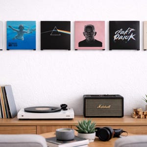 May include: A collection of vinyl record album covers displayed on a white wall. A wooden console table below holds a turntable, speaker, and decorative items. The album covers showcase diverse artists and album artwork.