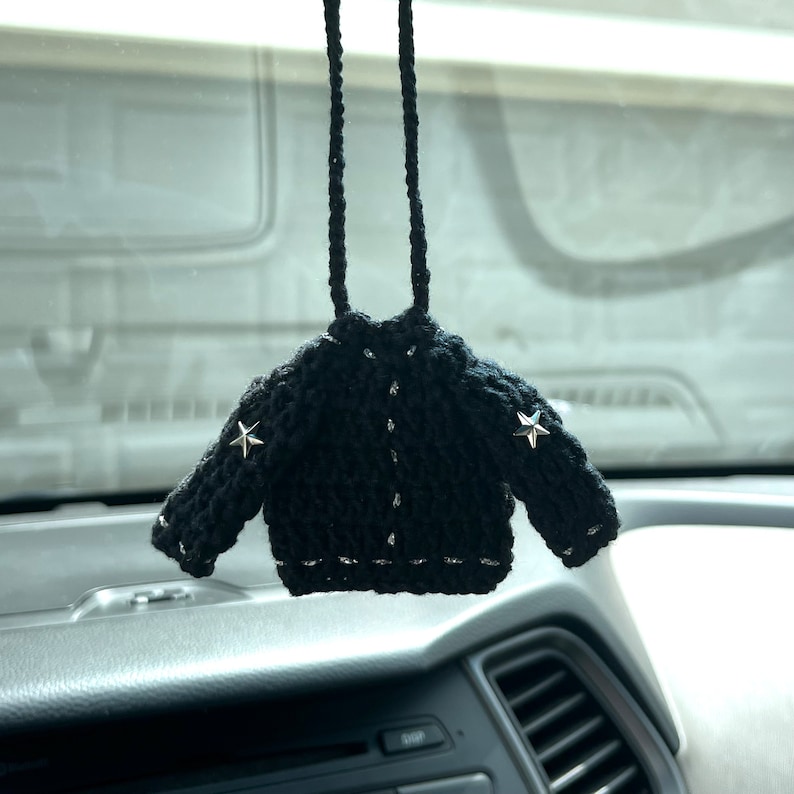 May include: A black, crocheted miniature jacket with silver star accents on the shoulders and silver trim. The jacket is hanging from a black cord, designed as a car accessory.