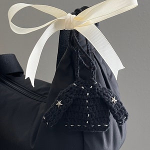 May include: A black bag with a cream-colored ribbon tied in a bow. A small, black crocheted jacket charm with silver accents and star-shaped embellishments hangs from the bag.