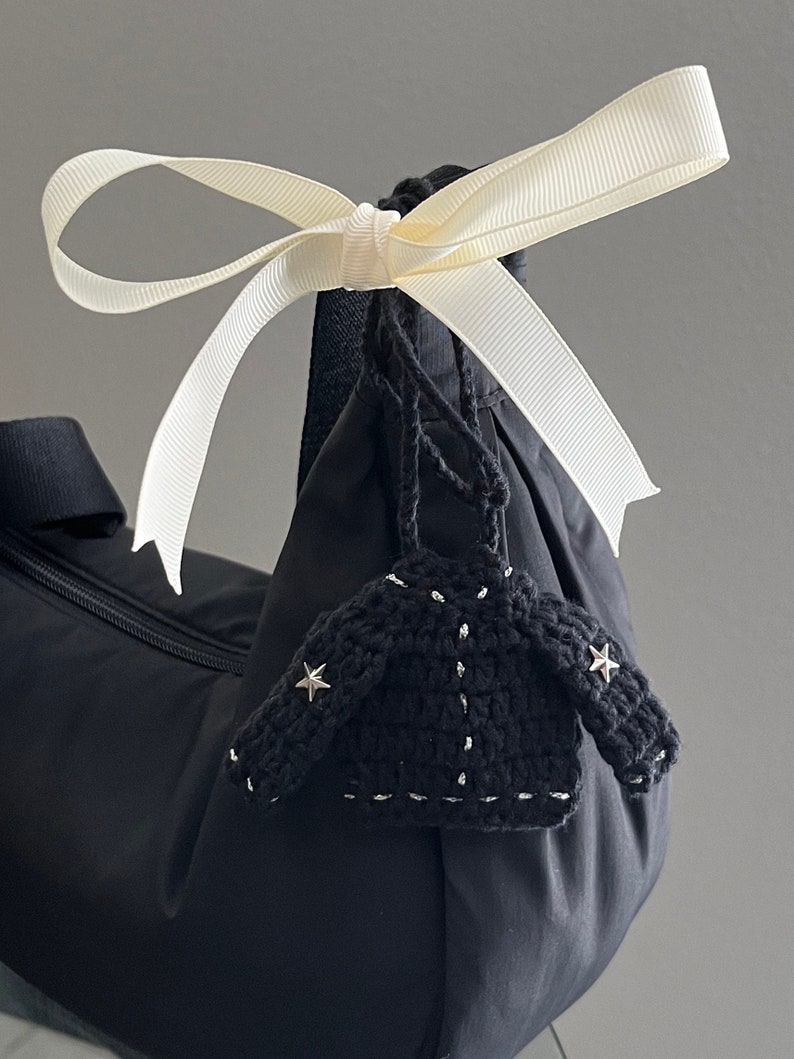 May include: A black bag with a cream-colored ribbon tied in a bow. A small, black crocheted jacket charm with silver accents and star-shaped embellishments hangs from the bag.