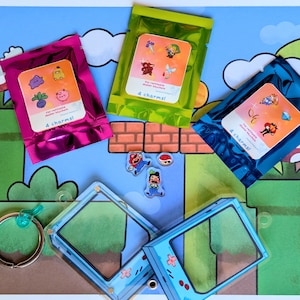 Może przedstawiać: Cztery kolorowe saszetki z napisem "4 charms" na każdej, zawierające małe zawieszki. Saszetki są ułożone na zielonym i niebieskim tle z motywem gry wideo. Są dwa przezroczyste breloki z plastiku w niebieskim i zielonym kolorze, przypominające konsolę do gier.