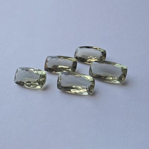 May include: Five loose, cushion-cut gemstones with a pale, translucent green hue. Each stone features a faceted surface that reflects light, creating a sparkling appearance. The gems are arranged in a cluster on a plain, light-coloured surface.