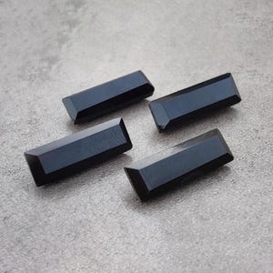 May include: Four rectangular, faceted black gemstones are displayed on a gray surface. Each stone has a polished, reflective surface and a geometric shape. The gemstones are arranged in a slightly offset pattern.