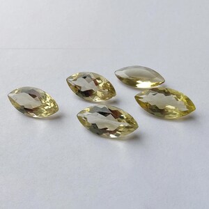 May include: Five marquise-cut gemstones with a pale yellow hue are arranged on a white surface. The faceted stones reflect light, showcasing their clarity and shape. The gems are similar in size and appear to be of high quality.