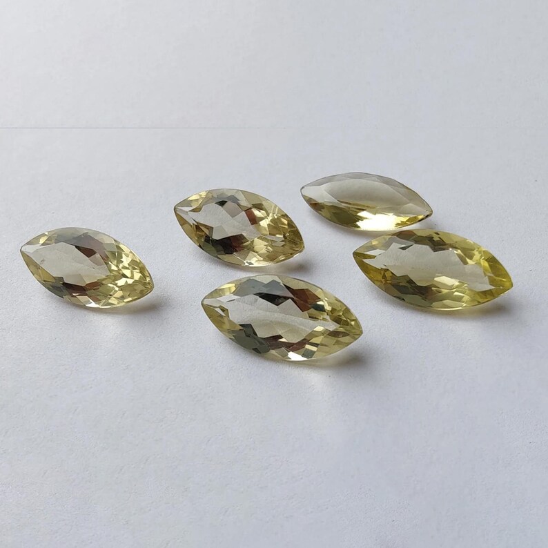May include: Five marquise-cut gemstones with a pale yellow hue are arranged on a white surface. The faceted stones reflect light, showcasing their clarity and shape. The gems are similar in size and appear to be of high quality.