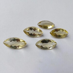 May include: Five faceted, marquise-cut gemstones in a pale yellow hue. The gems are arranged on a white surface, each displaying internal facets that catch the light. The stones are similar in size and shape, suggesting they are from the same source.