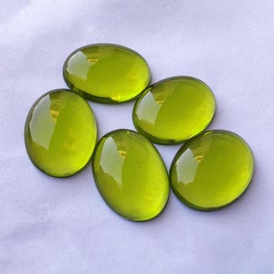 Może przedstawiać: Pięć owalnych, półprzezroczystych kamieni szlachetnych w kolorze chartreuse. Wypolerowane kamienie mają gładką, błyszczącą powierzchnię, odbijającą światło. Kamienie szlachetne ułożone są na białej powierzchni. Kamienie te mogą być używane do tworzenia biżuterii lub innych rękodzieł.